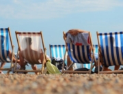 People relax in striped deck chairs facing the sea on a sunny pebble beach, symbolizing leisure, summer holidays, and coastal relaxation.